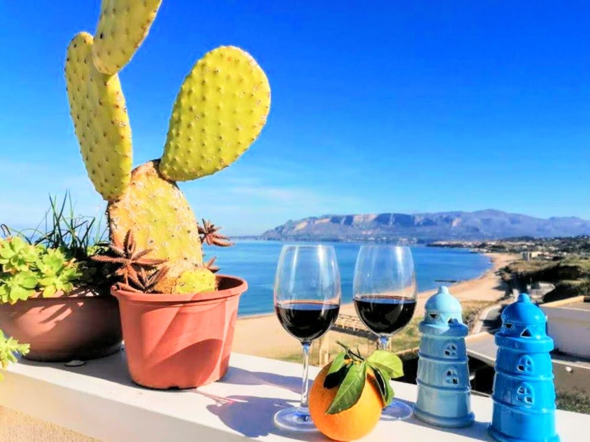 Um cacto, dois copos de vinho tinto, uma planta em vaso e um lanterna azul numa varanda com vista para o mar.