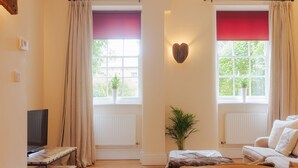 Un dormitorio con dos ventanas, un radiador, un televisor, un sofá, una mesita y una planta.