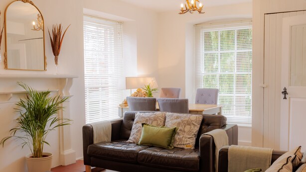 A living room with a brown leather sofa, a grey ottoman, and a dining table with chairs. There is a potted plant, a mirror, and a chandelier. - Game Cottage (Bridgnorth)