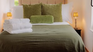A neatly made bed with green bedding and pillows, two bedside lamps, and a stack of white towels on the nightstand. - Game Cottage (Bridgnorth)