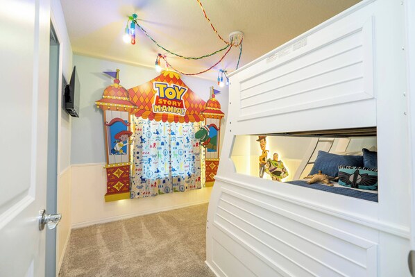 A child's playroom with a Toy Story Mania theme, featuring a bed with a view of the room, a wall-mounted TV, and a ceiling with string lights.