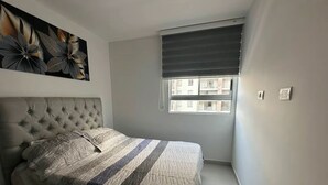 A bedroom with a tufted headboard, a striped bedspread, a window with a view of buildings, and a ceiling vent. - FURNISHED APARTMENT NEAR INTERNATIONAL HOSPITAL FOR RENT RENTAHOSUE (Floridablanca)
