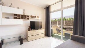 A modern living room with a TV, a glass coffee table, and a sofa. There is a large window with curtains and a view of a garden. - Home2Book Apart with Terrace & Pool in Costa Adeje (Adeje)