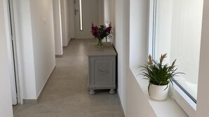 A narrow hallway with a light-colored tiled floor, a small side table with a vase of flowers, and a window with a potted plant. - Villa Reflets du Sud (LA GAUDE)