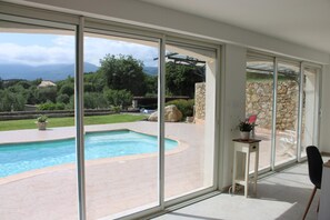 A modern home with a large glass door leading to a swimming pool, surrounded by a stone wall and a view of mountains. - Villa Reflets du Sud (LA GAUDE)