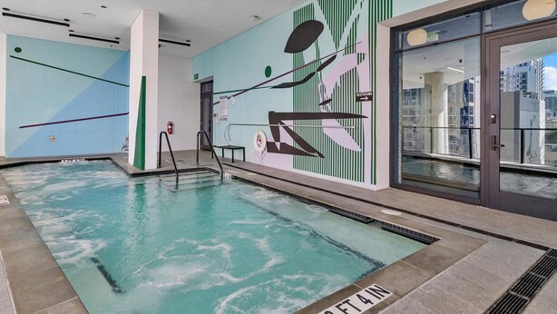 Ein Schwimmbecken mit Tiefenmarkierung von etwa 100 Zentimetern, einem Glastür-Eingang und einer Wand mit vertikalen, grünen Streifen sowie einem großen, abstrakten Design.