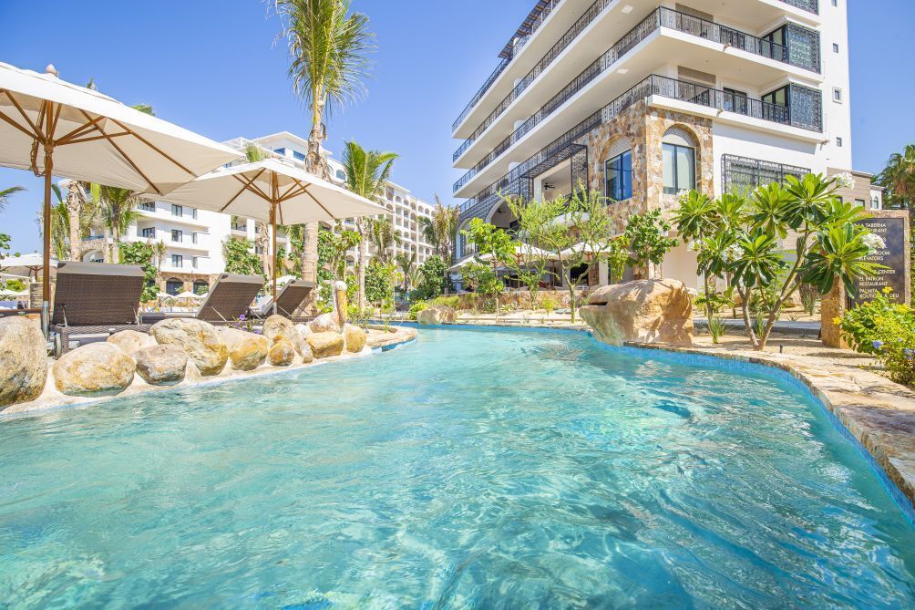 A pool area with lounge chairs and umbrellas, surrounded by palm trees and a multi-story building with balconies.
