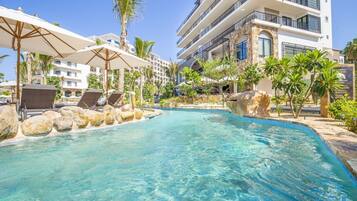 A pool area with lounge chairs and umbrellas, surrounded by palm trees and a multi-story building with balconies.