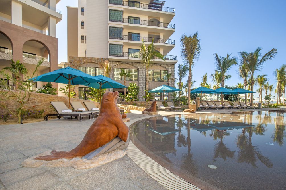 A resort pool area with lounge chairs and umbrellas, a large building with balconies, and palm trees.