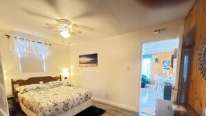 A bedroom with a bed, a ceiling fan, a painting, and a wooden floor. - The White Pine Cottage on the Cabot Trail (Englishtown)