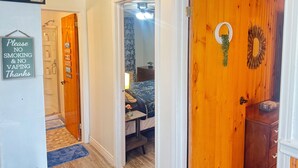 A hallway with a 'Please No Smoking & No Vaping Thanks' sign, a wooden door with a wreath, and a bed with a blue and white patterned bedspread. - The White Pine Cottage on the Cabot Trail (Englishtown)