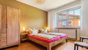 Un dormitorio con dos camas de madera, un armario, un ventanal con cortinas y una mesita.