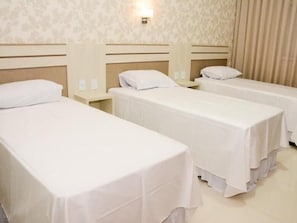 Ein Hotelzimmer mit drei Einzelbetten, jeweils mit weißer Bettdecke und passenden Kissen. Zwischen den Betten befindet sich ein Nachttisch mit einer Lampe, und die Wände sind mit einem dezenten Blumenmuster verziert.