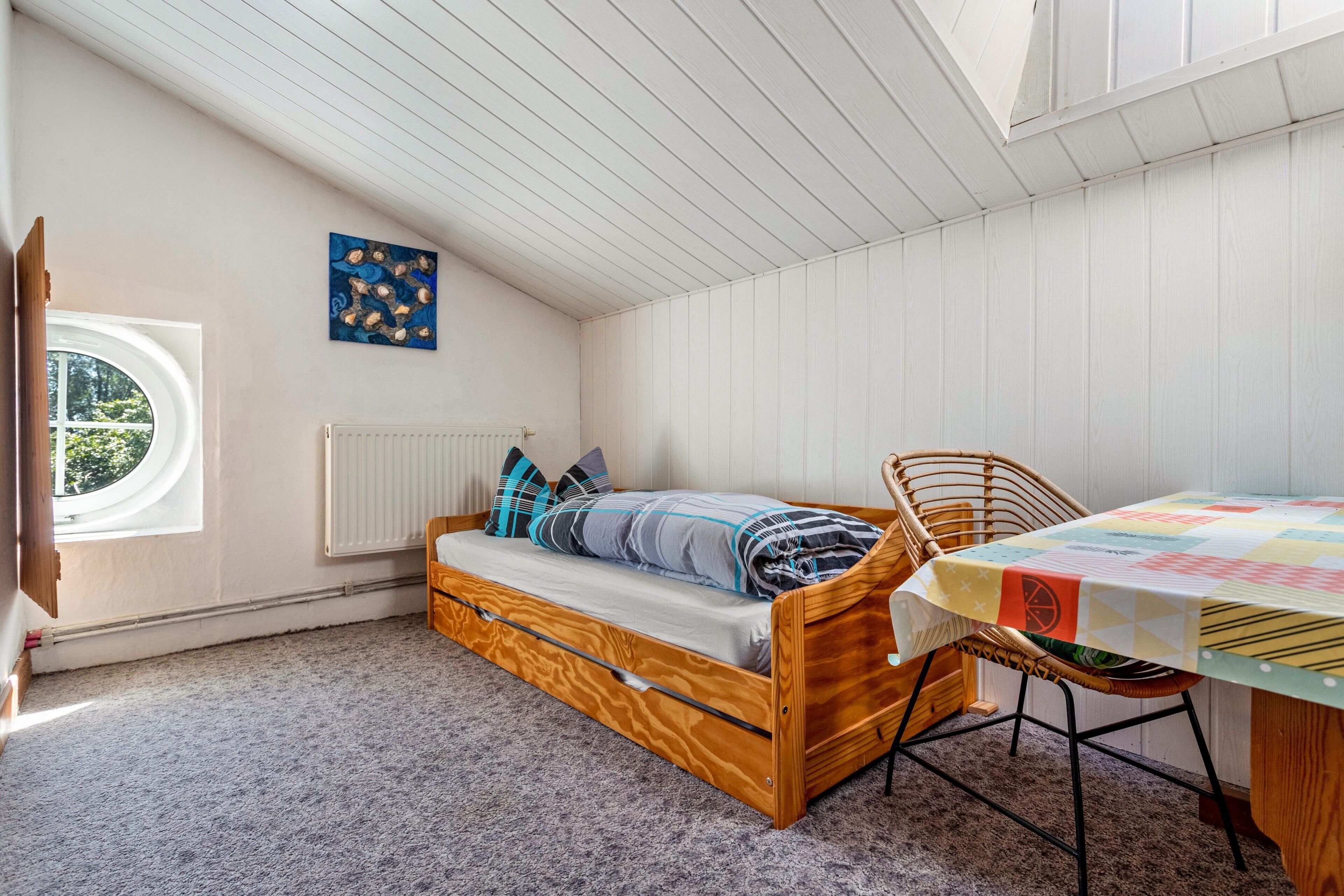 A room with a loft bed, a window, a radiator, a table with a tablecloth, and a wall art piece.