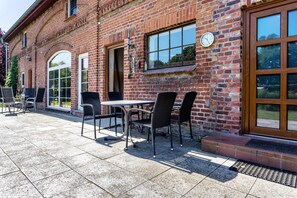 A brick building with a patio area featuring a table and chairs, a clock, and a large window. - Apartment 'Kranichblick' with Private Garden, Balcony, and Wi-Fi (Lüdershagen)