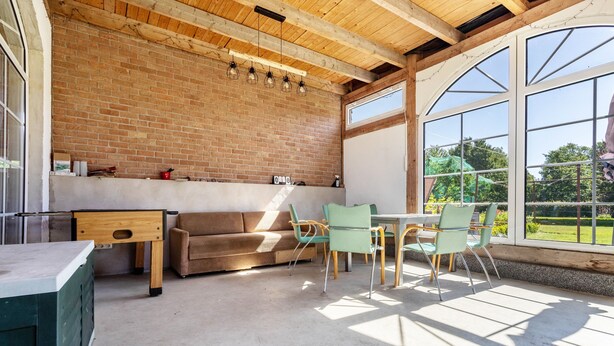 A room with a brick wall, wooden ceiling beams, a sofa, a table with chairs, and large windows.