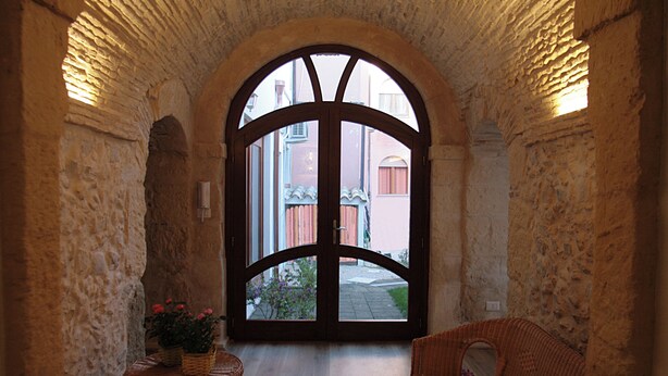Un pasillo con pared de piedra, suelo de madera, muebles de mimbre y un amplio arco que da al exterior.