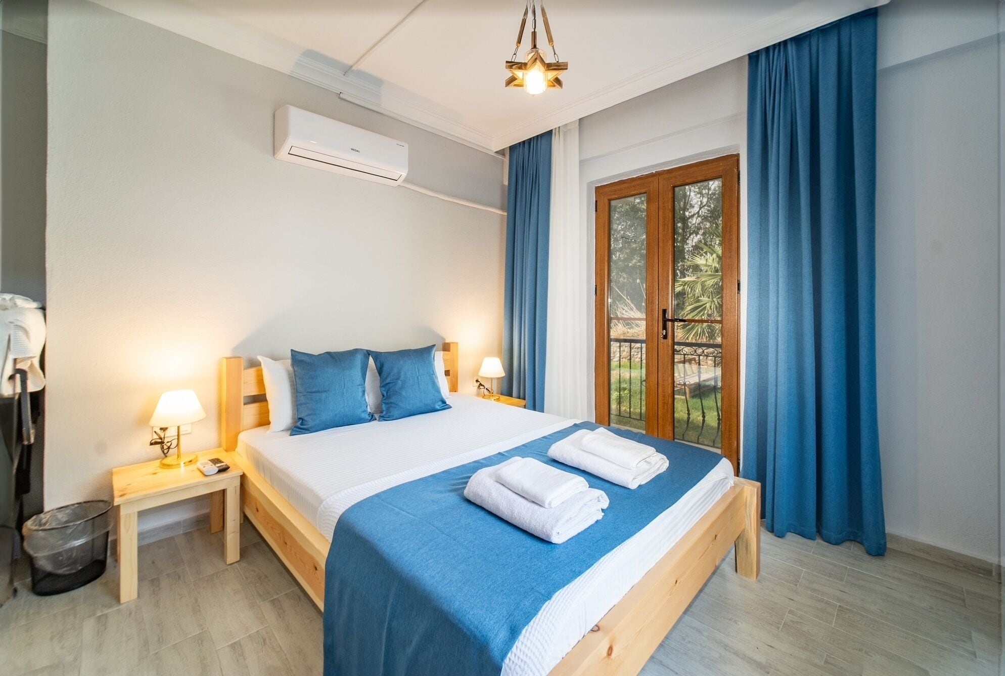 Quarto de hotel com cama, mesa de cabeceira, aparelho de ar condicionado, janela com cortinas e vista para o verde lá fora.