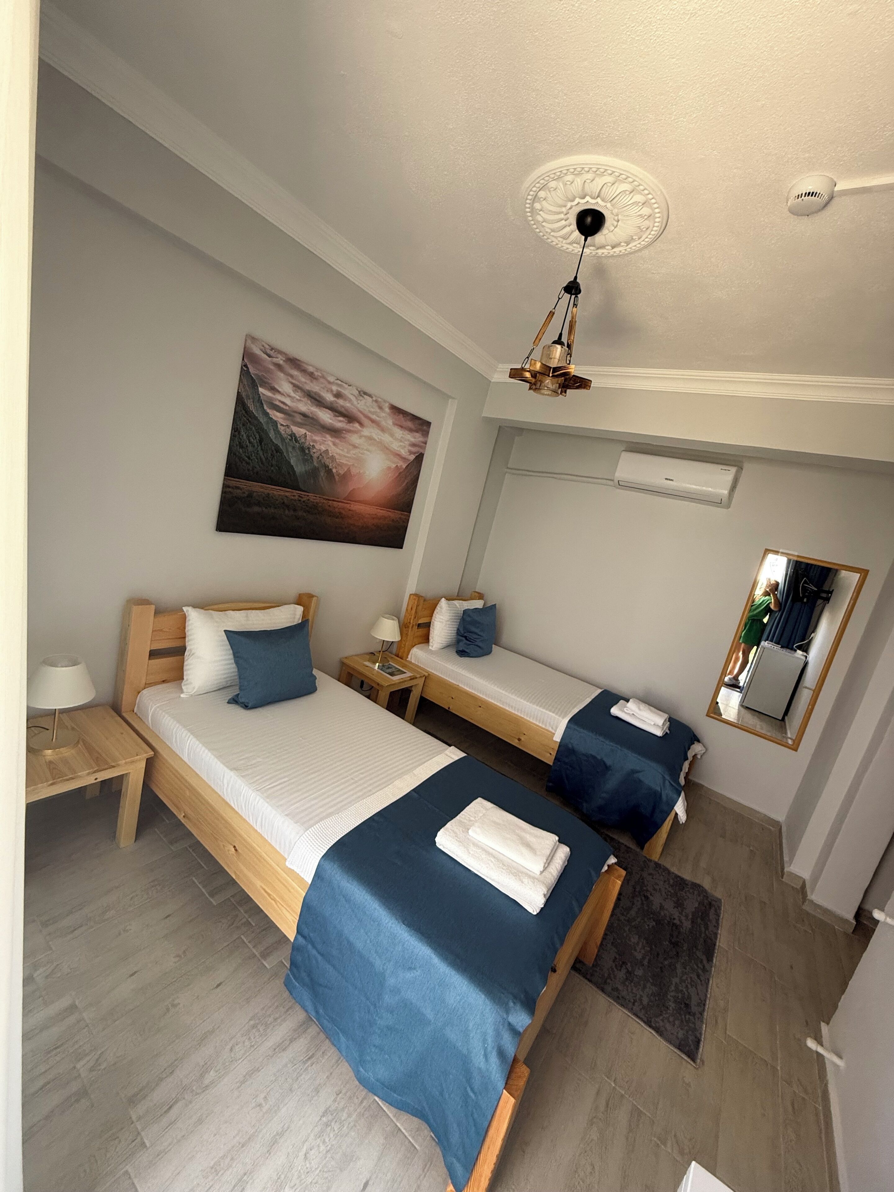 Habitación de hotel con dos camas individuales, ventilador de techo, aire acondicionado, un cuadro en la pared y un espejo.