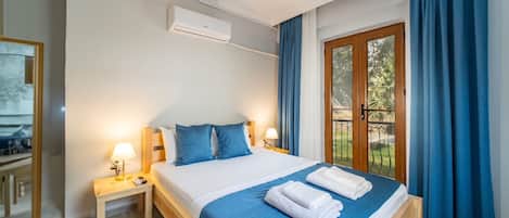 A hotel room with a bed, two bedside tables, an air conditioning unit, a window with curtains, and a view of the outdoors.