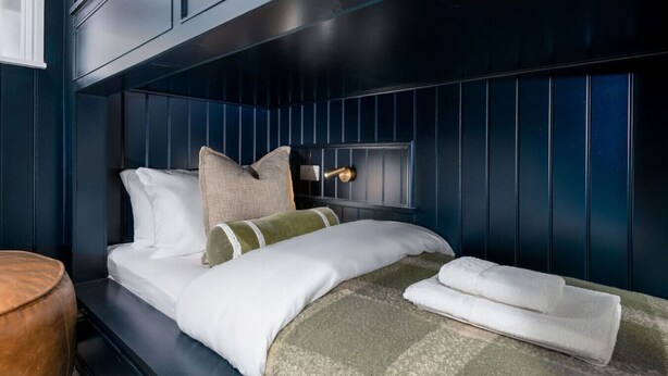 Une chambre à coucher aux couleurs sombres, avec un lit, des oreillers, une table de chevet et une lampe.