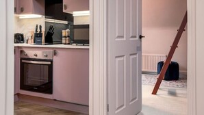 A modern kitchen with pink cabinets, a microwave, and a built-in oven. A wooden herringbone floor and a white door lead to a bedroom with a bed and a suitcase. - The Nevis Suite at Munro Guest House (Stirling)