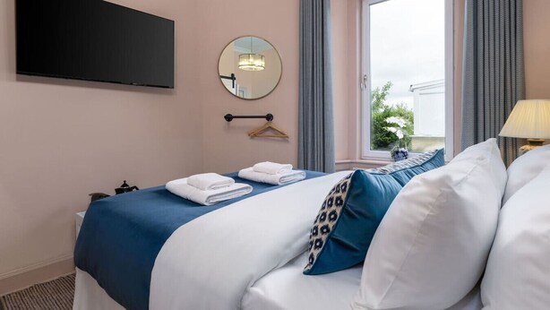 Habitación de hotel con cama, televisor de pantalla plana montado en la pared, espejo redondo y ventana con cortinas.