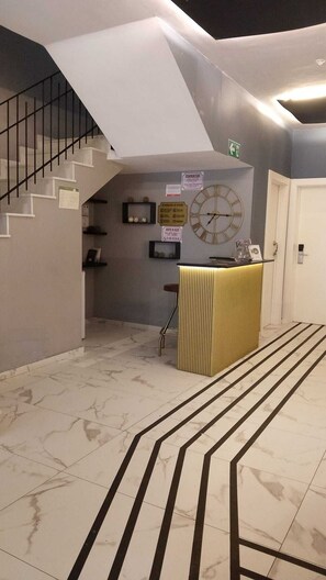 Un hall d’hôtel avec un escalier, un comptoir d’accueil, une horloge murale et un sol carrelé.