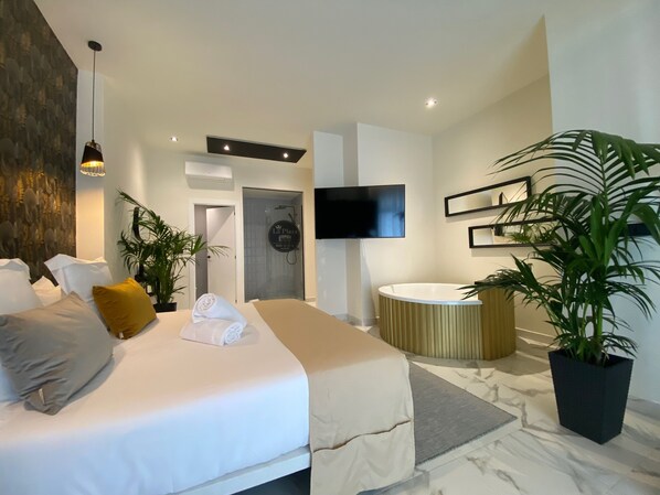 Une chambre d’hôtel moderne avec un grand lit, une baignoire ronde, un téléviseur fixé au mur et plusieurs plantes en pot.