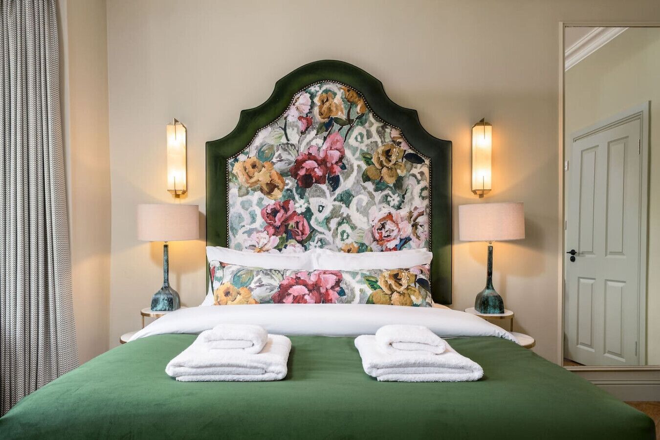 Une chambre d’hôtel avec un lit, une couvre-lit verte, une tête de lit à motifs floraux, deux lampes de chevet et une porte.
