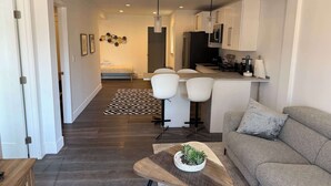 A modern living room with a sofa, a coffee table with a plant, and a dining area with chairs and a kitchen in the background. - Cozy 2-Bedroom Guesthouse with Private Patio (San Francisco)