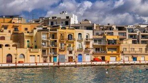 A coastal cityscape with multi-story buildings, a waterfront promenade, and a clear sky. - Charming home Apt with private balcony in B`Bugia (Birżebbuġa)