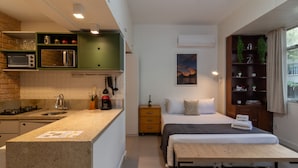 Una habitación de hotel compacta con kitchenette, una cama con mesita de noche, una pequeña mesita, una mesilla auxiliar y un estante con objetos decorativos.