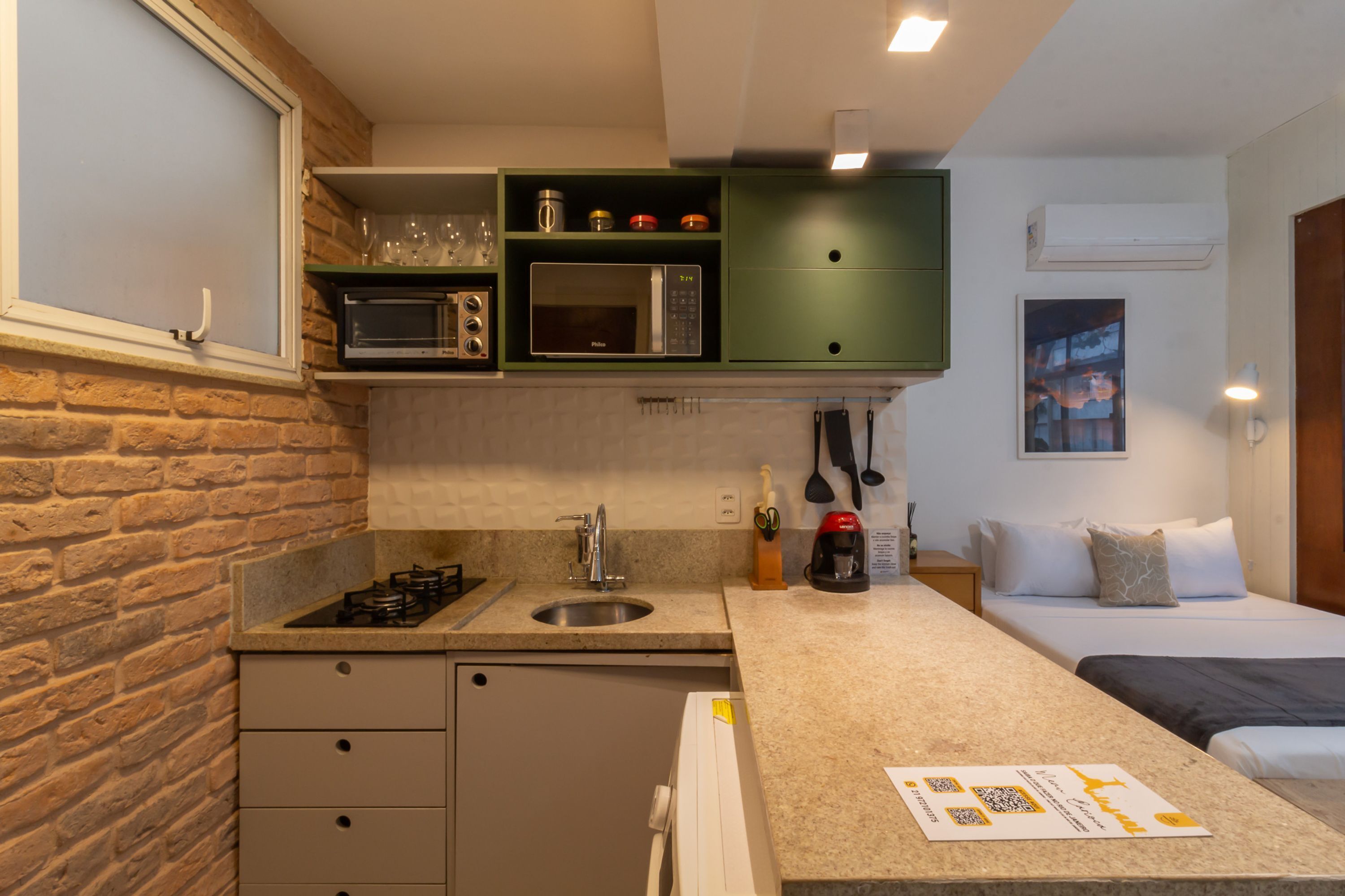 Cocina compacta con lavamanos, microondas y armarios verdes. Se ve una cama con almohadas en la habitación contigua.