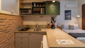 Cocina compacta con lavamanos, microondas y armarios verdes. Se ve una cama con almohadas en la habitación contigua.
