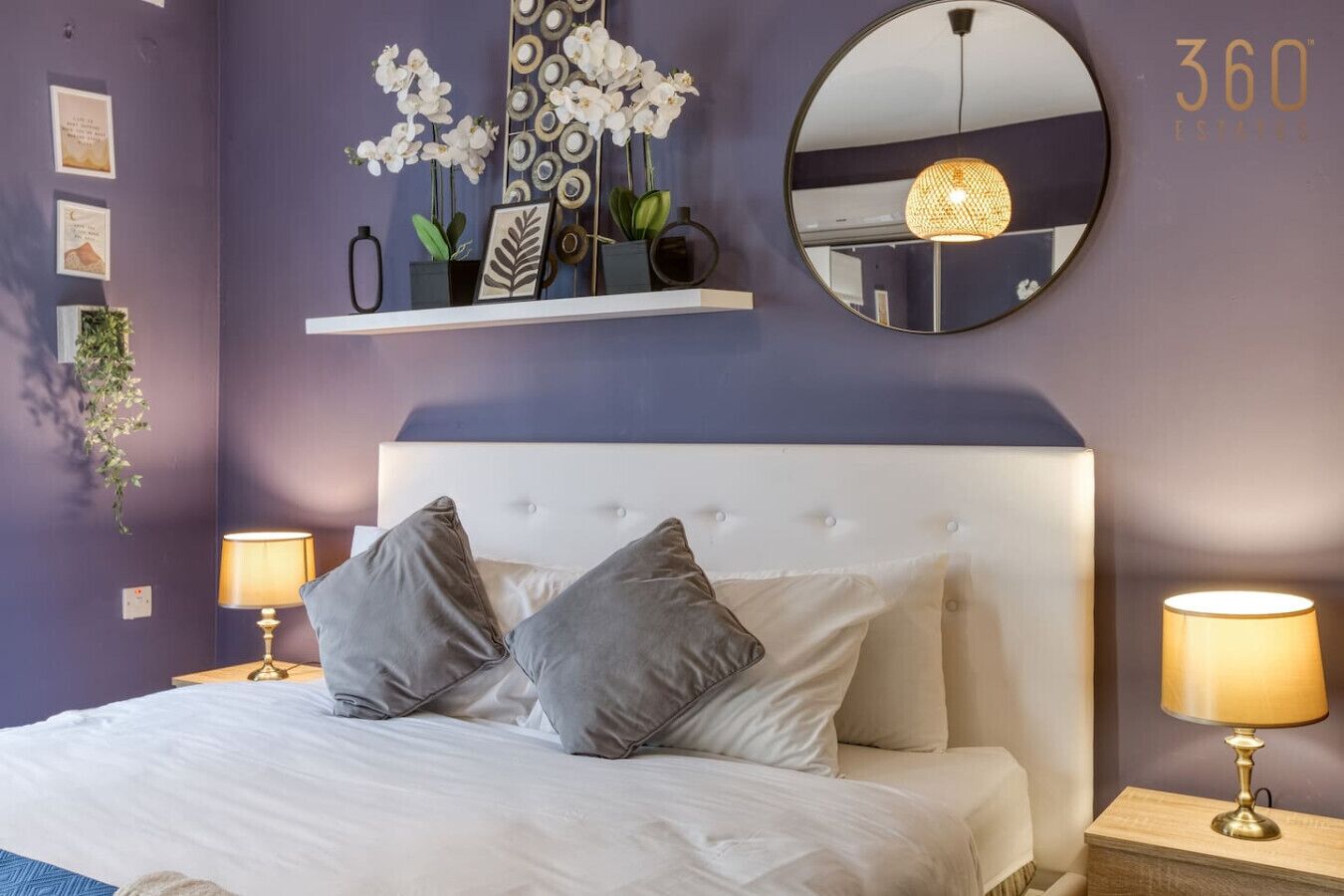 A bedroom with a white headboard, two bedside tables with lamps, a round mirror, and decorative items on the wall.