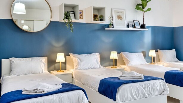 Habitación de hotel con tres camas, ropa de cama blanca, detalles en azul, un espejo redondo y un estante con adornos.