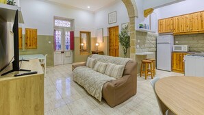 A cozy living room with a sofa, a TV, and a dining table. There is a kitchen area with wooden cabinets and a refrigerator. - Maltese Maisonette in Historic Isla with Sea Views (L-Isla)