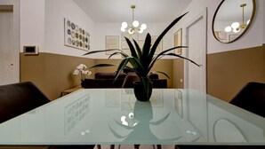 Un comedor con mesa de vidrio, dos sillas, una planta en maceta, un sofá, un espejo montado en la pared y una lámpara en el techo.