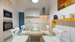 A compact kitchen with built-in appliances, a dining area with a glass table and white chairs, and a wooden shelf with kitchen items. - Cozy 3rd-Floor Valletta Apartment by the Seafront (Il-Belt Valletta)