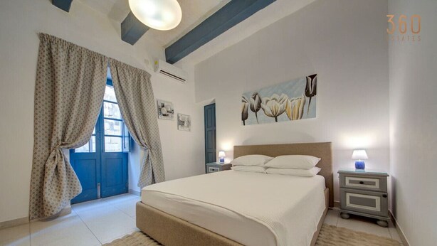 A bedroom with a large bed, two bedside tables with lamps, a window with curtains, and a painting on the wall. - Cozy 3rd-Floor Valletta Apartment by the Seafront (Il-Belt Valletta)