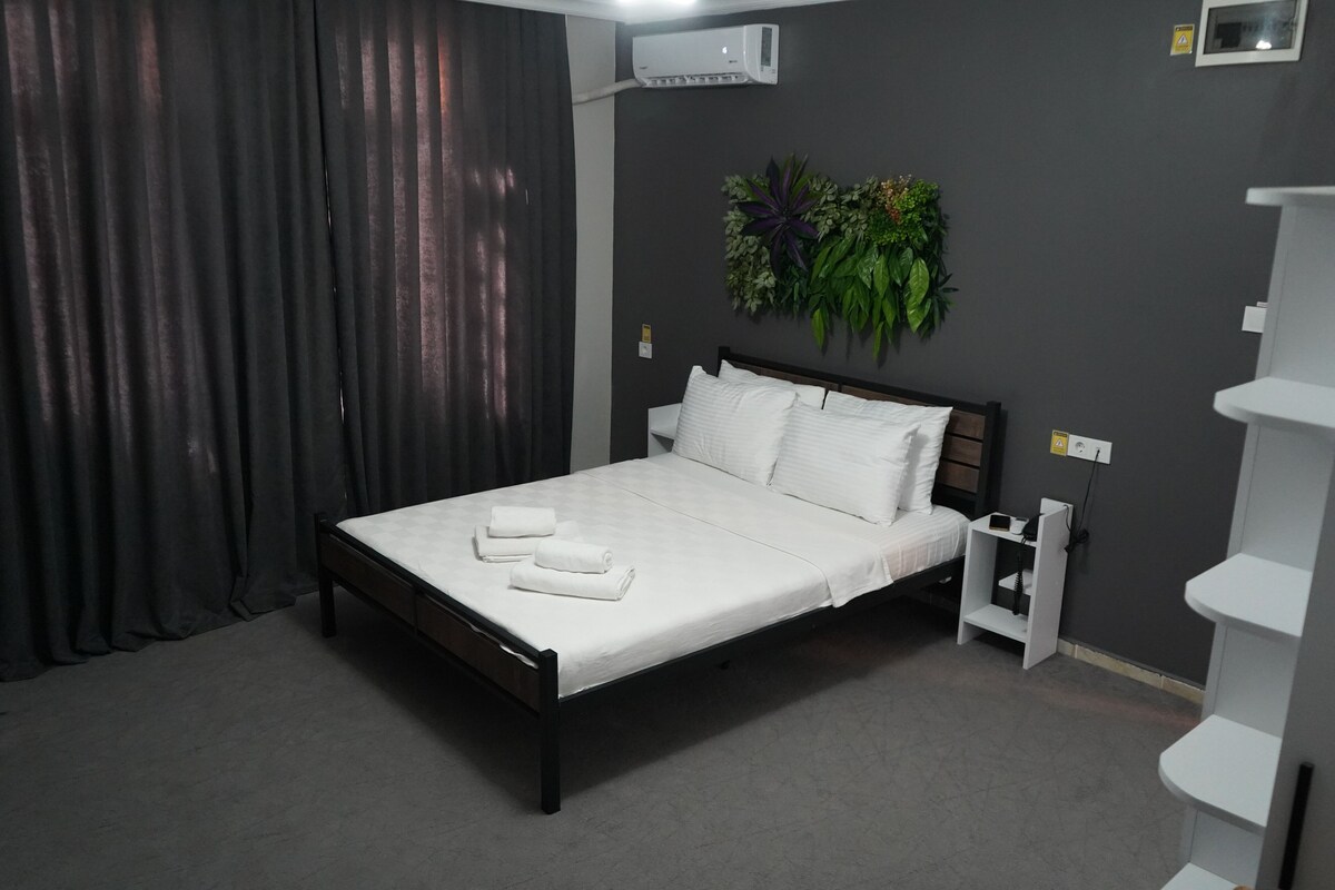 Ein ordentlich bezogenes Bett mit weißen Leinen und Handtüchern, eine dunkelgraue Wand mit einer Zimmerpflanze und ein weißer Nachttisch mit Steckdose.