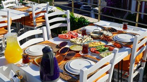 Ein Tisch ist für eine Mahlzeit gedeckt, mit verschiedenen Gerichten, einem Orangensaftkrug und einem Teekessel auf einer Terrasse mit weißen Stühlen und einem Fliesenboden.