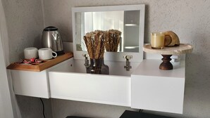 Ein modernes Badezimmer mit weißem Waschtisch, einem Spiegel, einer Kaffeemaschine, einer Vase mit Trockenblumen und einer kleinen Ablagefläche mit Kerze und Dekorationsobjekt.