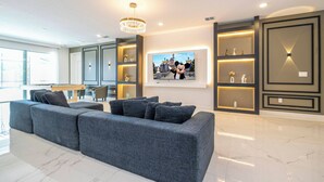 Uma sala de estar moderna com um sofá modular cinza, uma área de jantar com mesa de madeira e uma cozinha ao fundo.