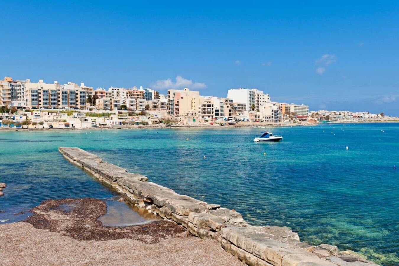 A coastal cityscape with a clear blue sea, a stone pier, and a boat.