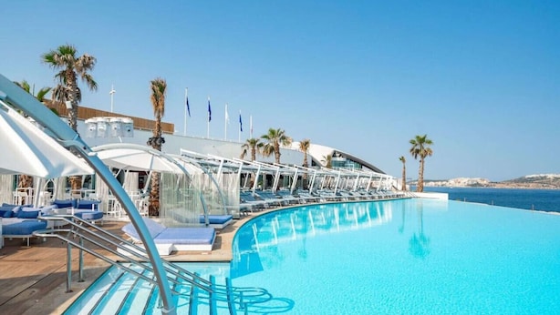 A cruise ship deck with a large pool, sun loungers, and palm trees.