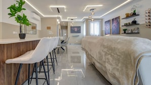 A modern hotel room with a large bed, a dining area with bar stools, a kitchen area, and a living space with a sofa and decorative plants.