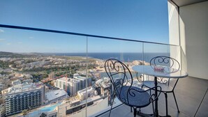 Ein Balkon mit Glasgeländer, der auf eine Stadtlandschaft und das Meer blickt. Zwei Metallstühle und ein kleiner runder Tisch sind vorhanden.