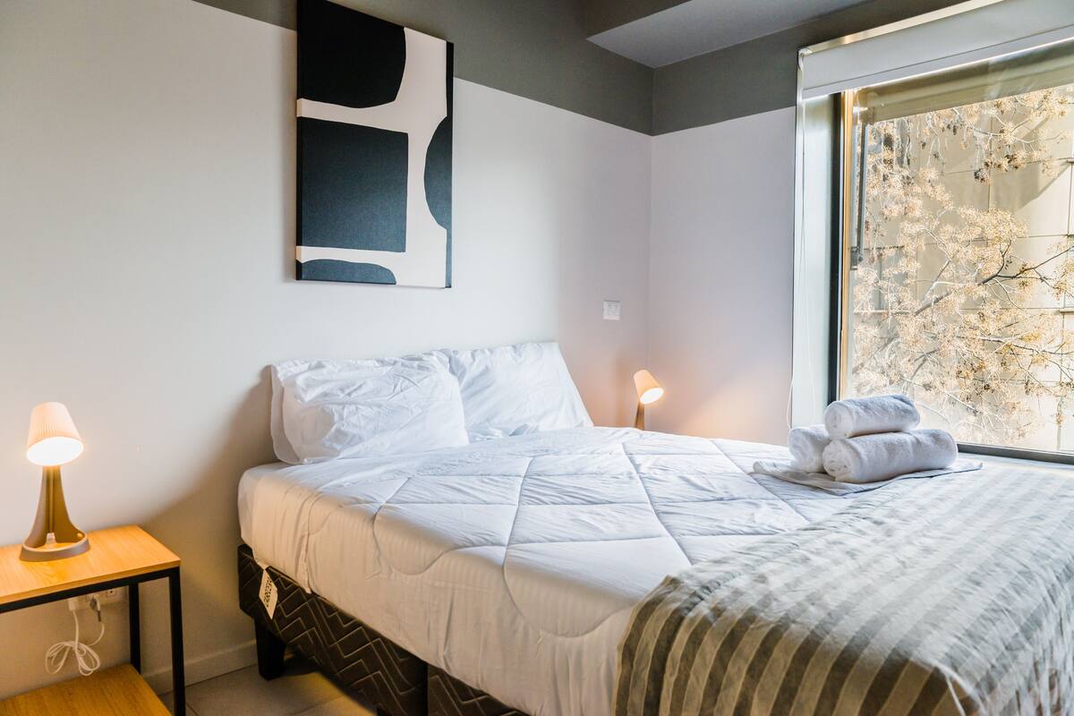 A neatly made bed with white linens, a bedside lamp, and a window with a view of trees.